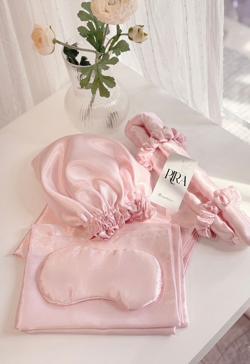RIRA satin sleep set in blush pink, including a bonnet, sleep mask, pillowcase, and scrunchies, styled on a white table with flowers.