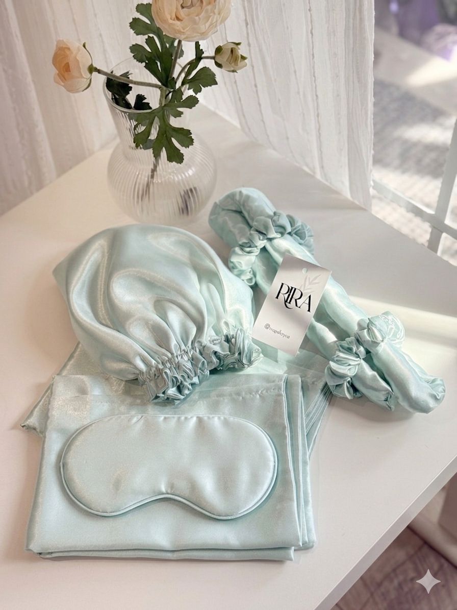 RIRA satin sleep set in mint green, including a bonnet, sleep mask, pillowcase, and scrunchies, styled on a white table with flowers.