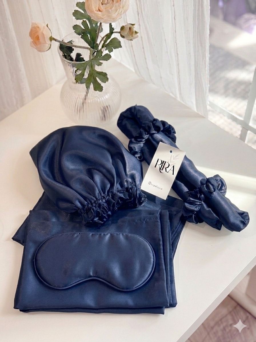 RIRA satin sleep set in navy blue, including a bonnet, sleep mask, pillowcase, and scrunchies, styled on a white table with flowers.